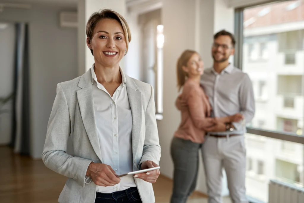 portrait-of-female-real-estate-agent-looking-at-the-camera-while-happy-couple-in-standing-in-the-background