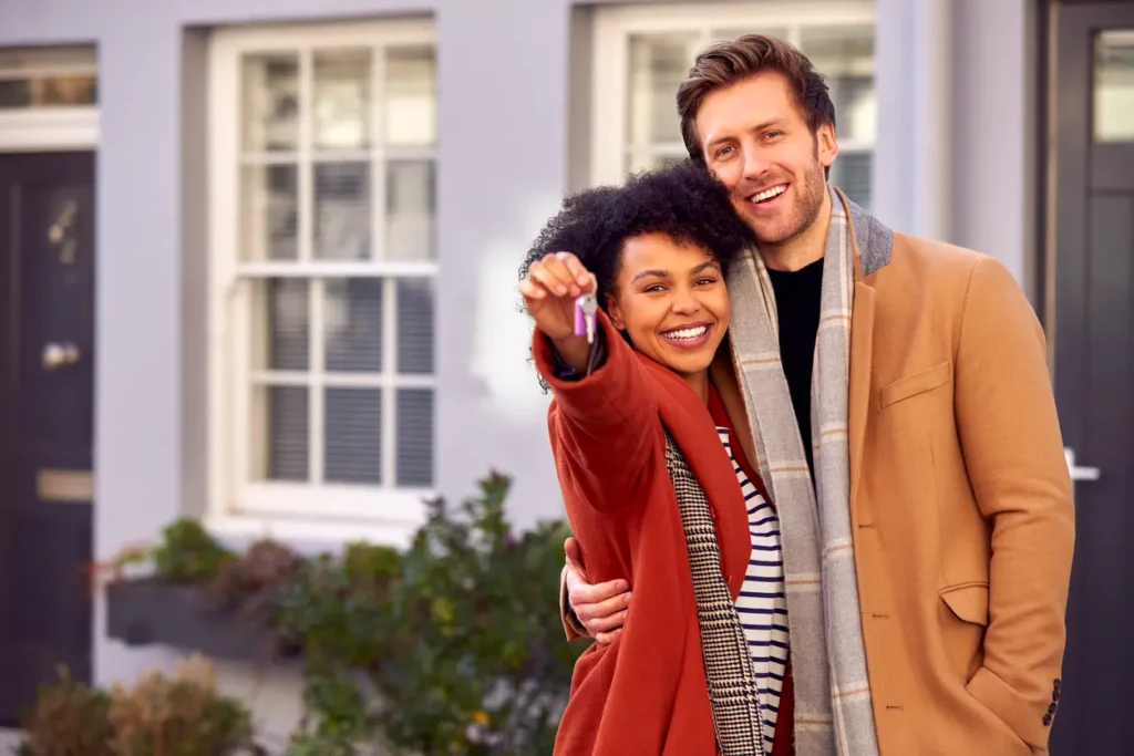 portrait-of-multi-cultural-couple-outdoors-on-moving-day-holding-keys-to-new-home-in-fall-or-winter