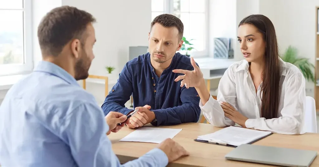 A couple sits with a property manager, asking questions about the lease agreement on the table.