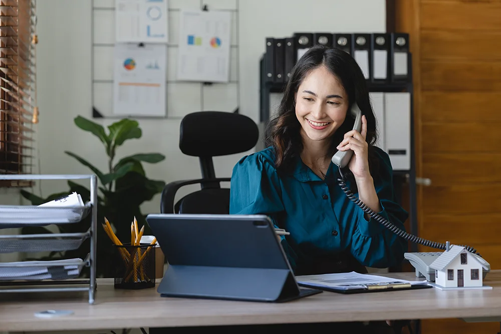 A woman sits at a desk with a laptop and forms, speaking on the phone.