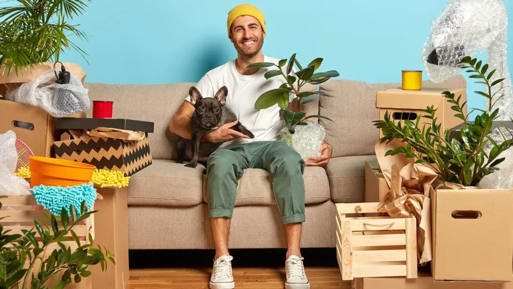 Indoor shot of delighted carefree house owner sits on couch and hugs pet.