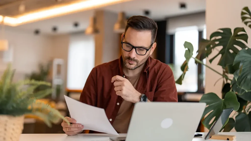 Serious millennial man using laptop sitting at the table in a home office, looking at the paper, communicating online, writing emails, distantly working or studying on computer at home