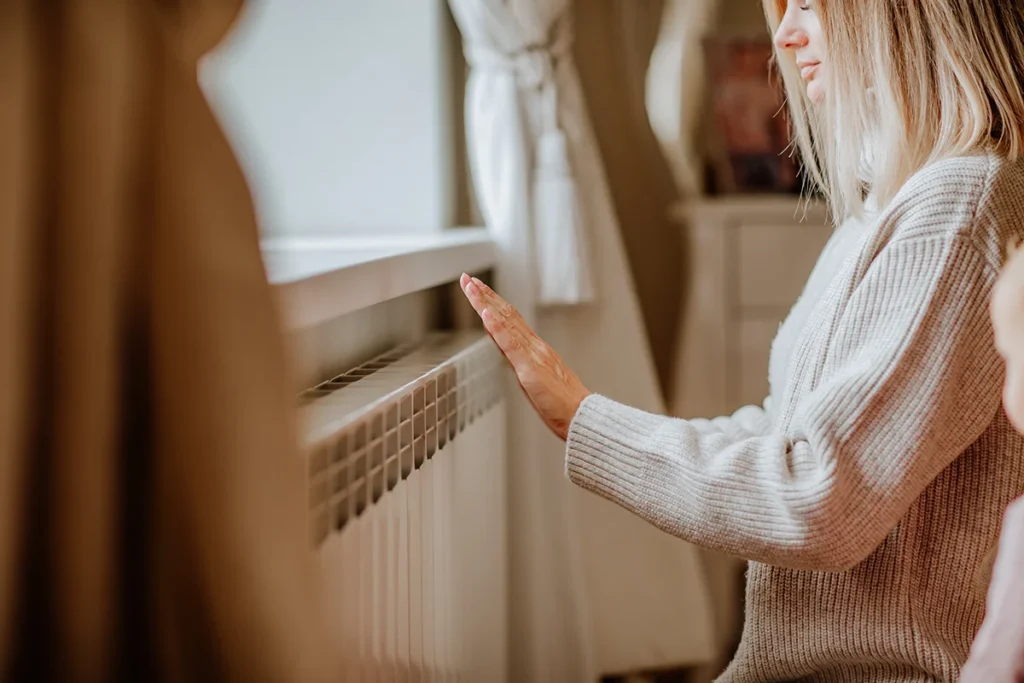 Young woman in long winter beige sweater is posing at home near the radiator.