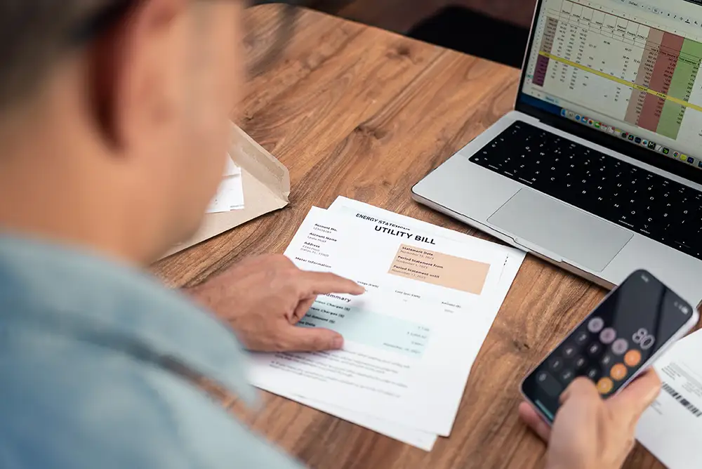 Man reviewing utility bills at home while using a smartphone calculator.