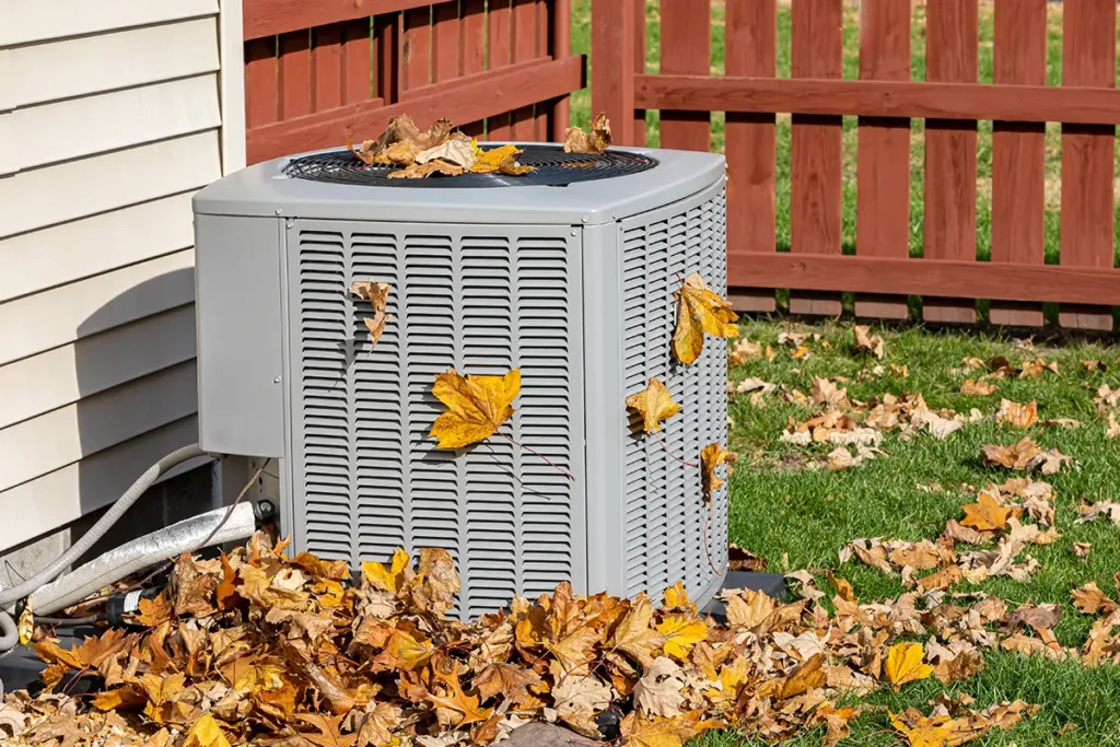An outdoor AC condenser surrounded by fallen leaves