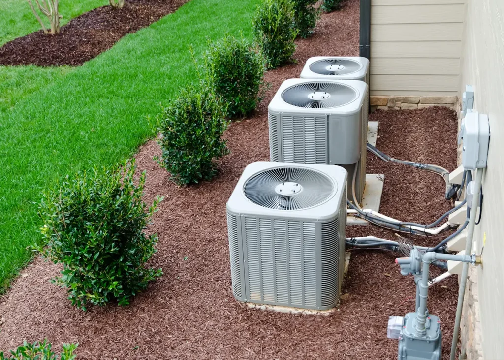 A row of three AC condensers outside a home, flanked by mulch, bushes, and grass